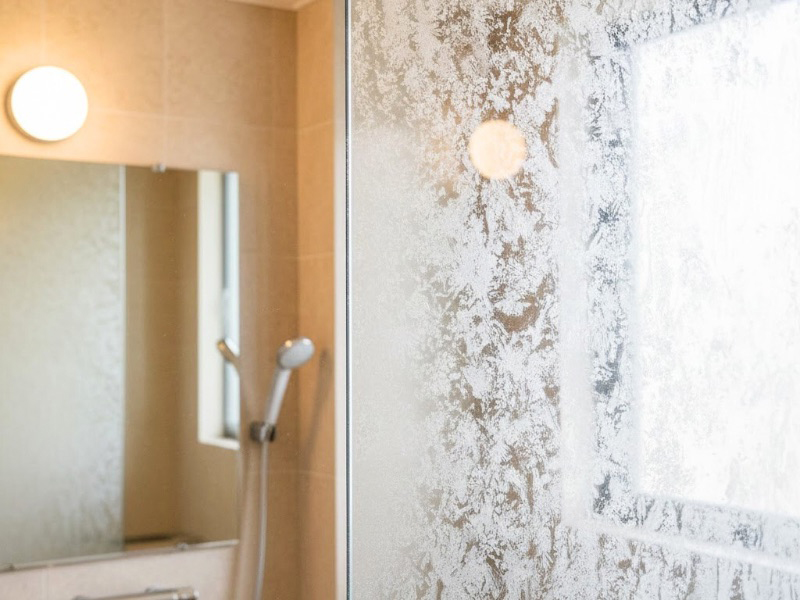 温泉旅館やホテルの大浴場の鏡とガラスが白く曇り、水垢やウロコ汚れで視界が悪くなっている様子。清掃では落としにくい固着汚れの問題を示すイメージ。